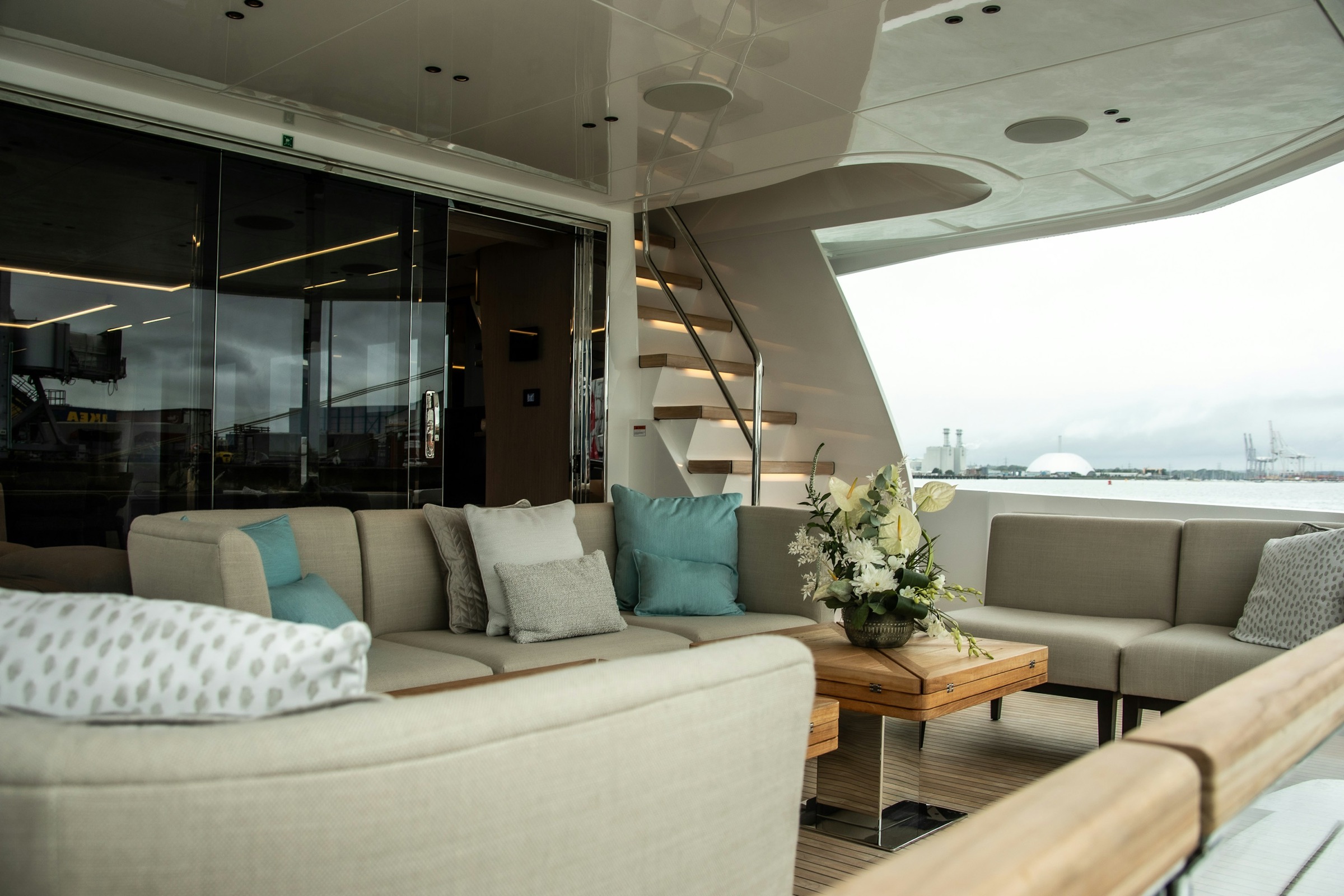 The aft saloon of a yacht in soft daylight, cream linen seating and teak decking opening onto a calm harbour beyond the rail.