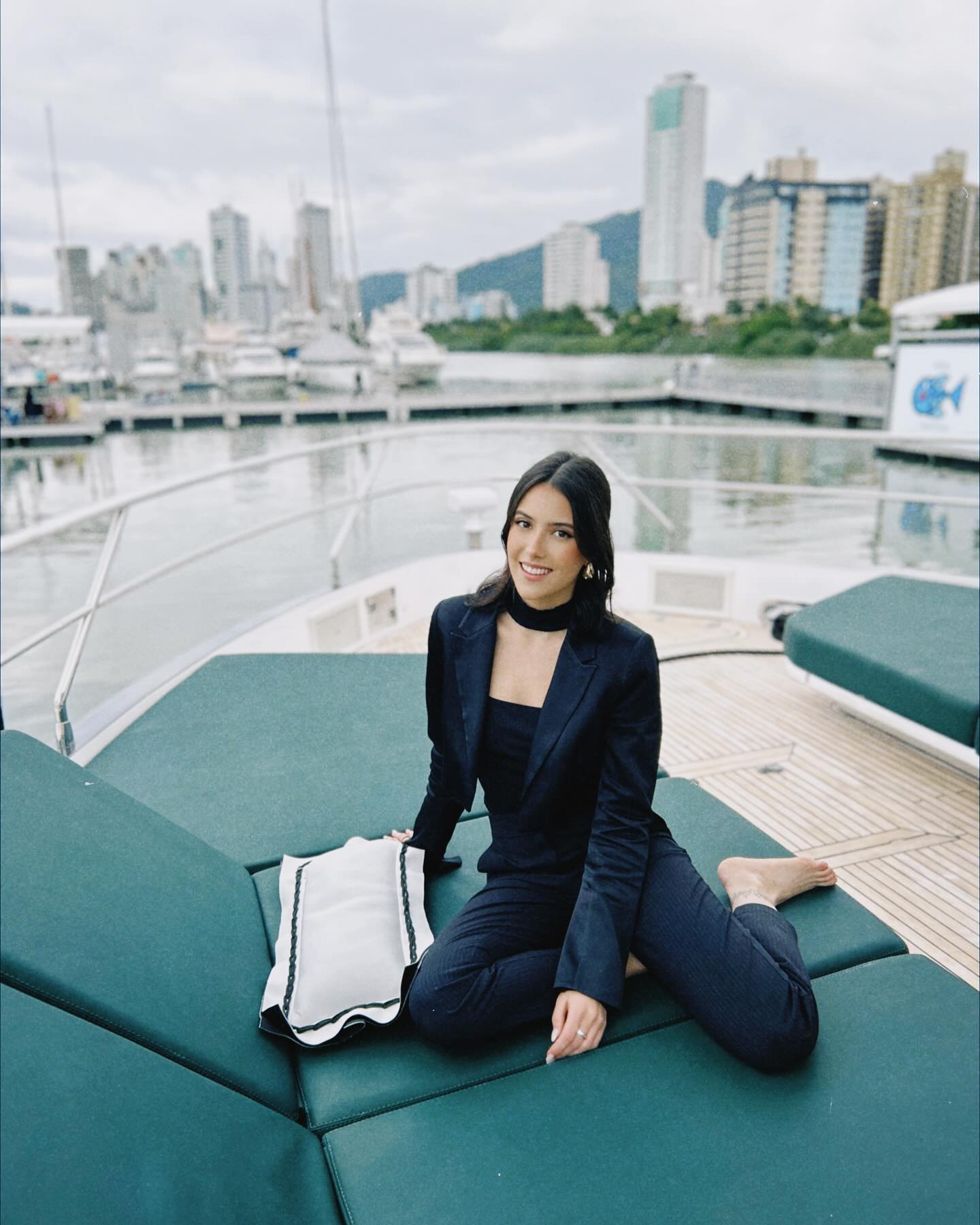 Olivia Larsen seated on the foredeck of a yacht in a navy suit, a marina and city skyline behind her.