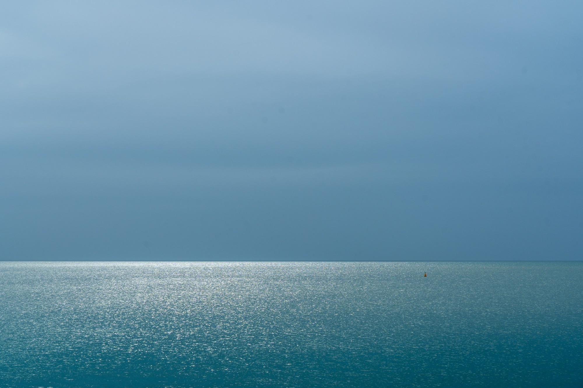 A still sea meeting a grey-blue sky at the horizon, a band of silvered light resting on the water.