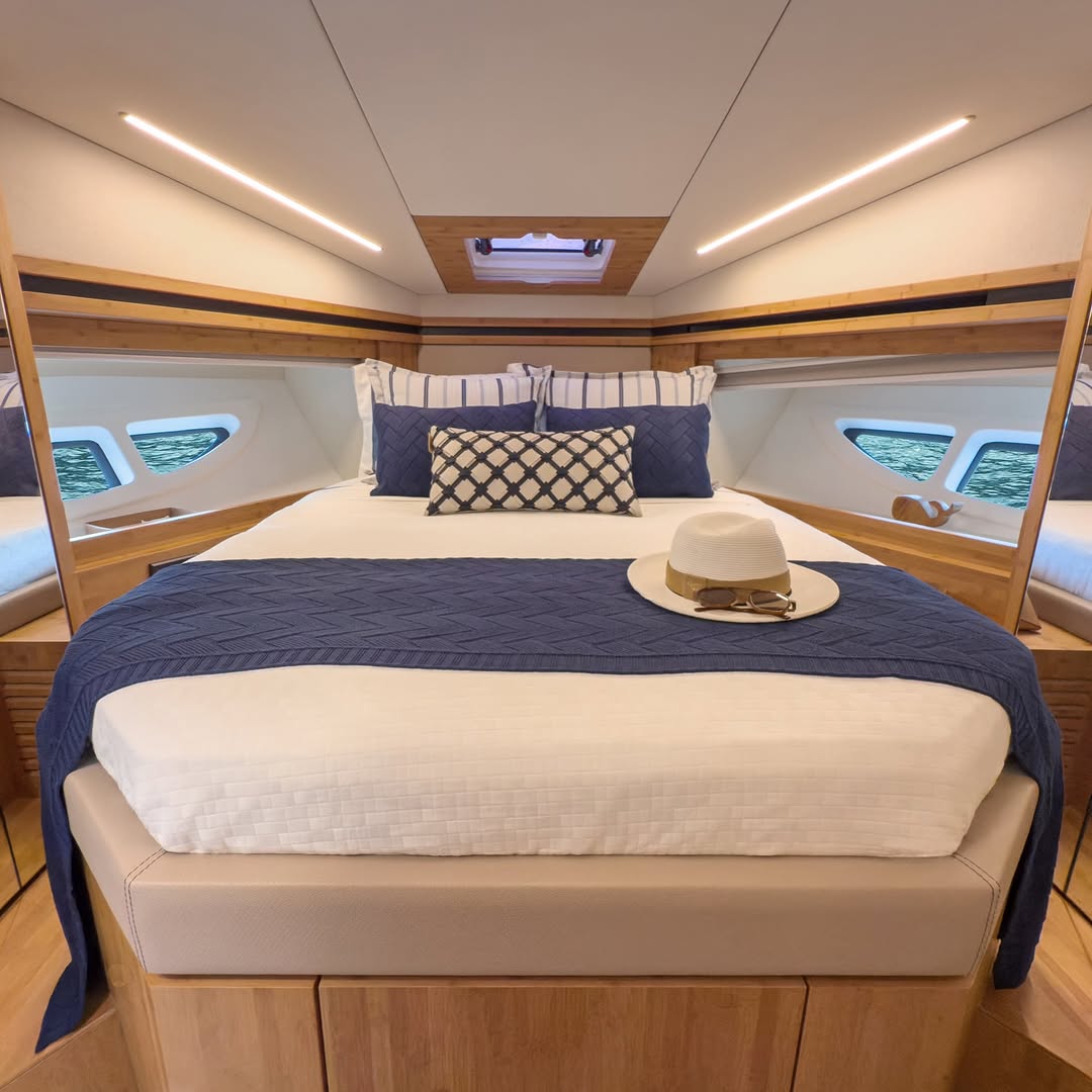 Owner's cabin aboard a yacht: a centred bed dressed in white with a navy knitted throw and a straw hat resting on it, the sea visible through hull windows.