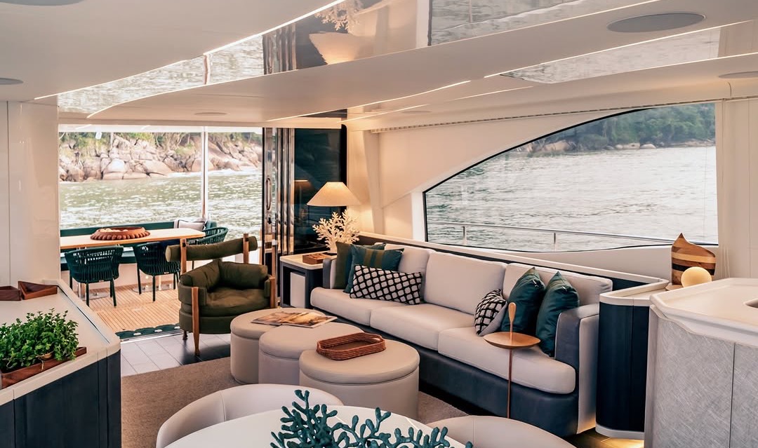 Main saloon of a 24-metre yacht: a cream sofa layered with green cushions in soft lamplight, a rocky shoreline seen through the open aft doors.