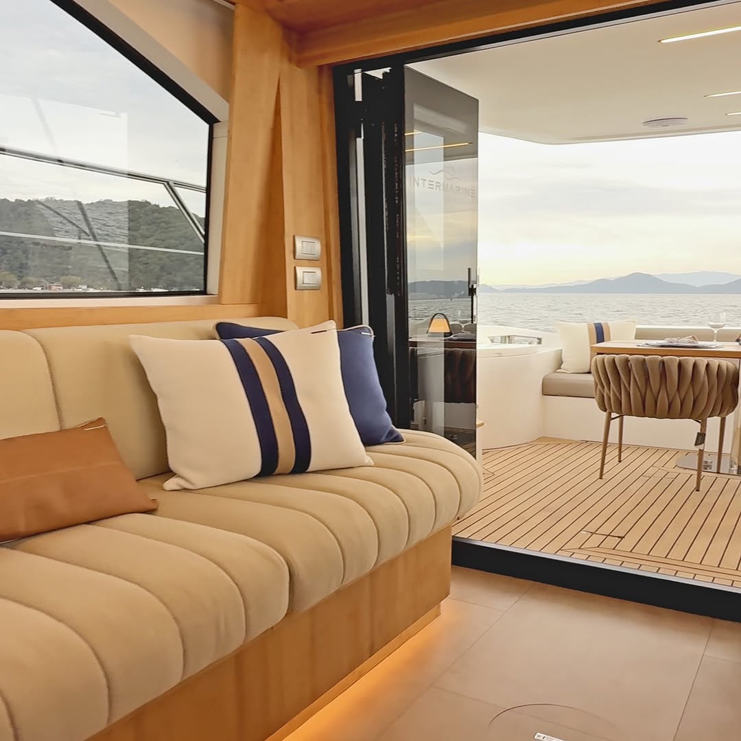 Yacht saloon at golden hour: a ribbed linen settee with striped cushions beside an open glass door leading to the teak aft deck and the sea.